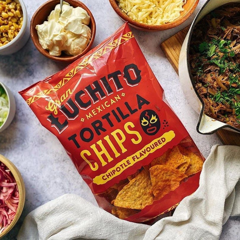 En pose med Gran Luchito Chipotle Tortilla Chips (150 g), omgitt av skåler med revet ost, rømme, mais, løk og stekt kjøtt på en kjøkkenbenk.
