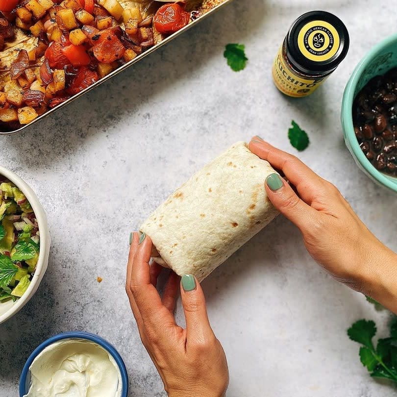 Hender som holder en innpakket burrito på en benk, omgitt av skåler med rømme, salat, svarte bønner, stekte grønnsaker og en krydderkrukke – alt klart for de perfekte hjemmelagde burritoene med Gran Luchito Burrito Wraps 360 g.
