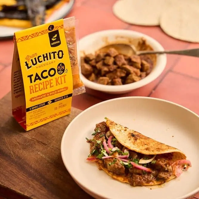 En tallerken med en fylt meksikansk taco står ved siden av en skål med langtidskokt kjøtt og et «Gran Luchito Birria Recipe Kit» på 300 g på et bord med tortillas i bakgrunnen.