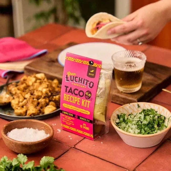 Et «Gran Luchito Carnitas»-oppskriftssett på 300 g, med ferskhakket koriander, salt og smakfull fylling av trevlet svinekjøtt, står på et bord. En person holder en meksikansk taco full av smak ved siden av et glass øl.