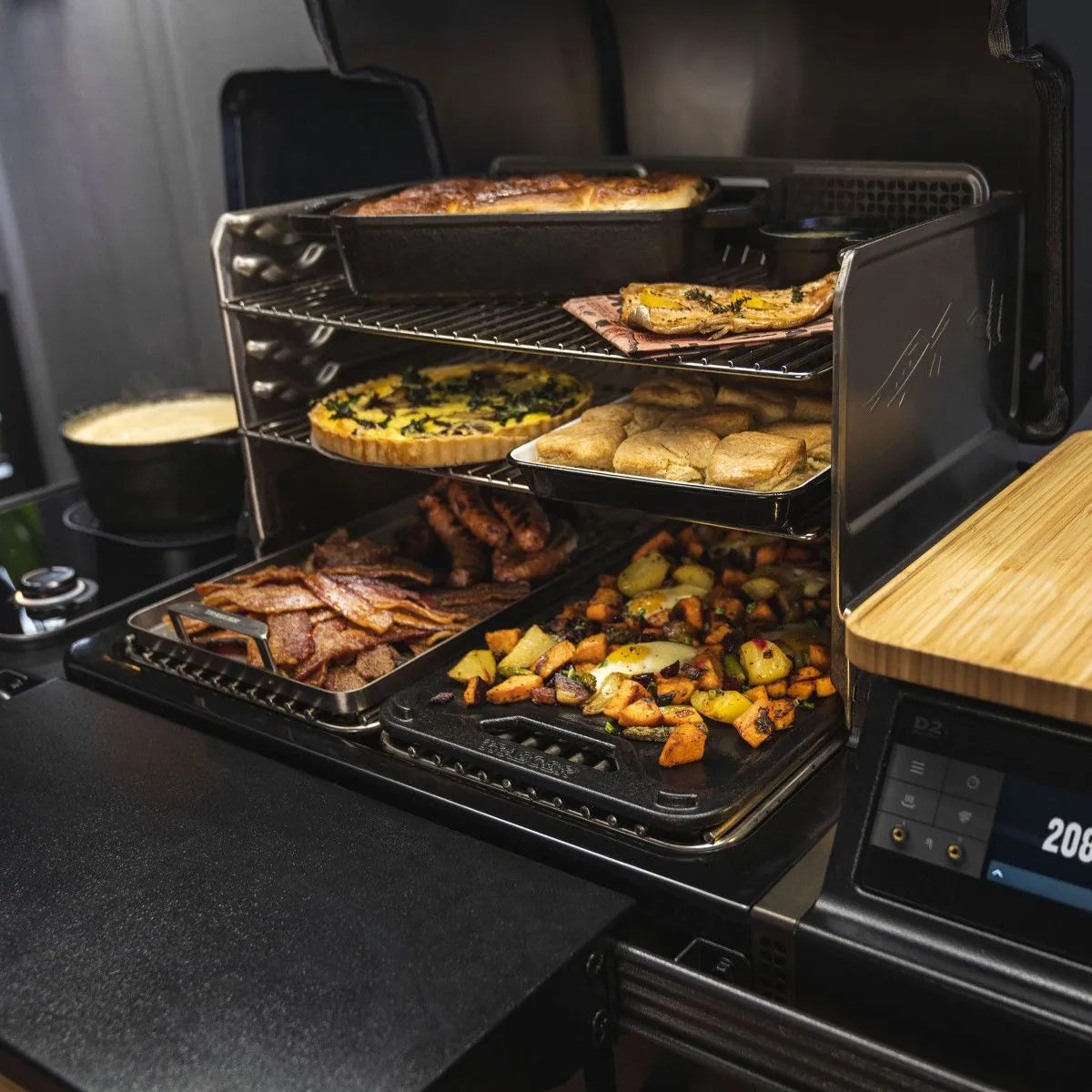 En Traeger Timberline L pelletsgrill fylt med stekte grønnsaker, bacon, rundstykker, quiche og gryterett gir liv til smakene fra utendørs matlaging - akkurat som å lage mat over en vedfyrt grill, med en gryte som putrer ved siden av.
