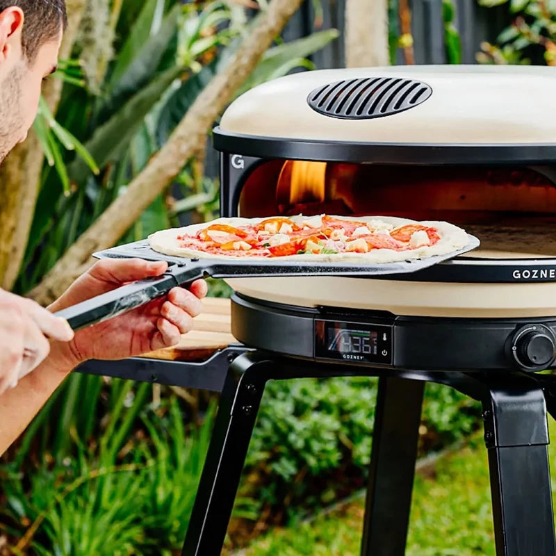 Noen bruker en Gozney Pro Pizzaspade 14" Large til å skyve en ubakt pizza inn i en utendørs pizzaovn som står på et stativ i en hage.