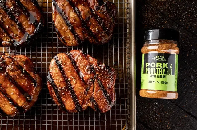 Fire grillede svinekoteletter på en rist ved siden av et glass Traeger Pork & Poultry Rub 200g fra Traeger, en smakfull grillkrydderblanding for svin og kylling med eple- og honningaroma.