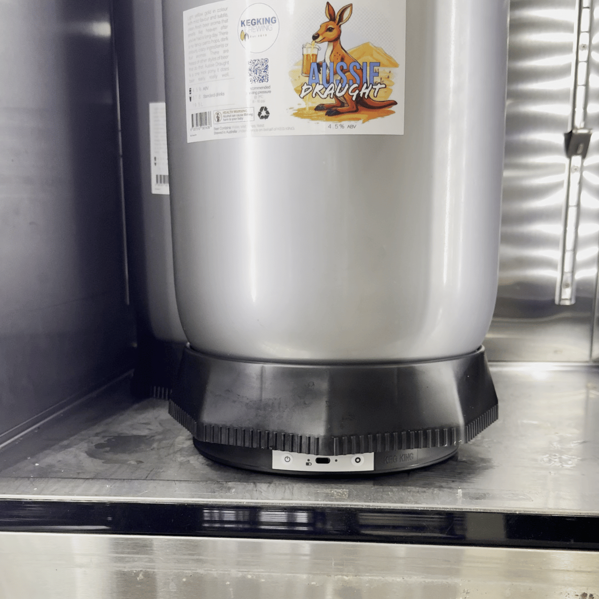 Et sølvfarget fat merket Aussie Draught står på en svart sokkel inne i et metallkjøleskap, utstyrt med Keg King KegMaster KS1 KegScale - vekt til corneliusfat for nøyaktig overvåking.
