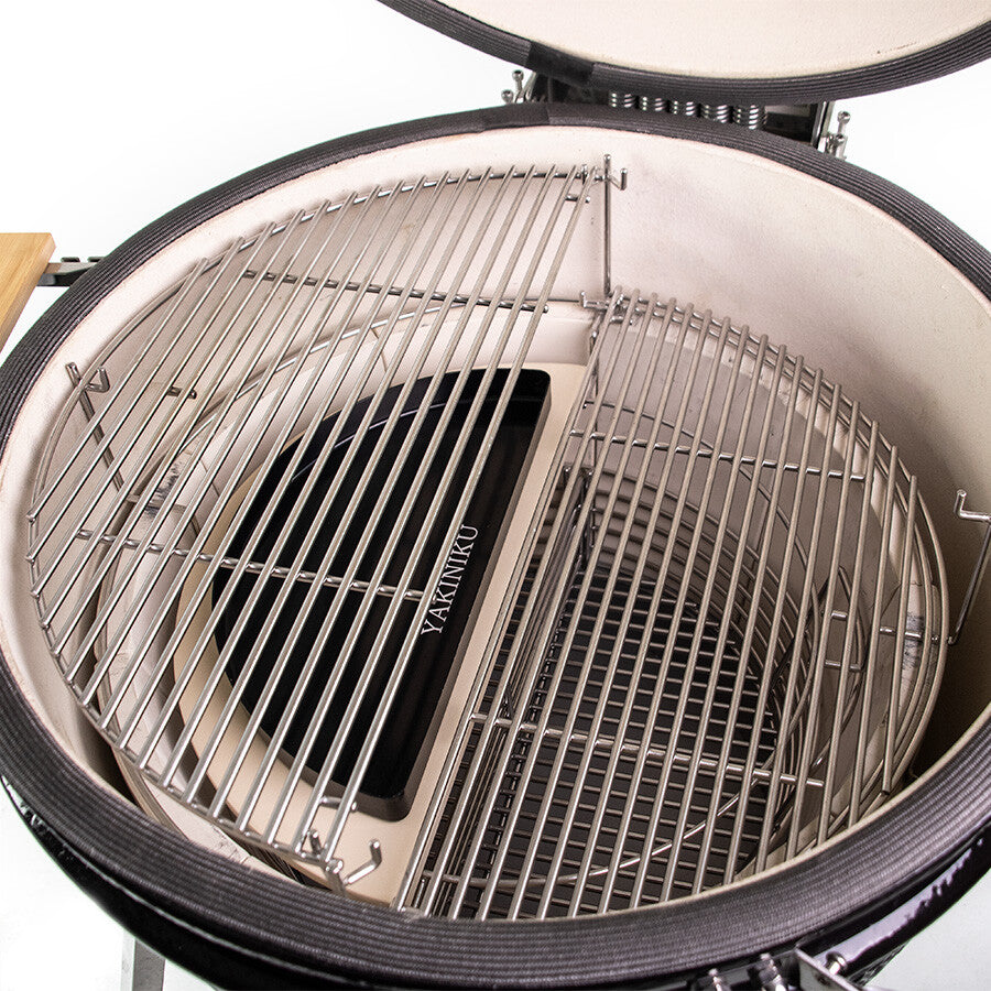 Den åpne kamadogrillen i keramikk har metallrister og et YAKINIKU Halvmåne Dryppfat XLARGE / LARGE for indirekte grilling. På innsiden er det en YAKINIKU-varmedemperplate, og grilllokket er delvis synlig på toppen.