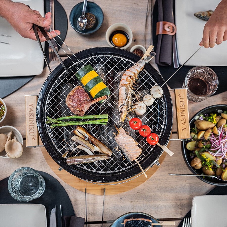 To personer griller ulike grønnsaker, sjømat og kjøtt på en YAKINIKU Round Shichirin Japansk Bordgrill, med tallerkener, drikke og kjøkkenutstyr på spisebordet.