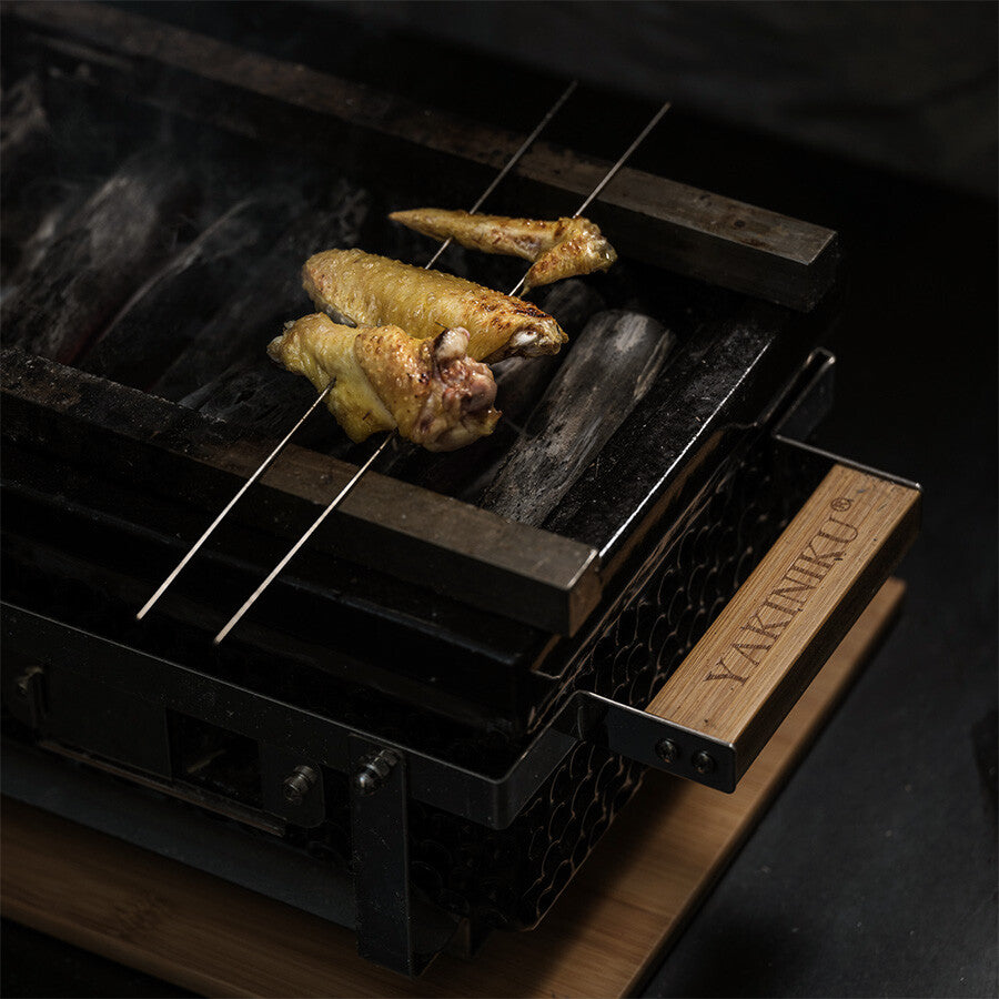 To metallspyd med grillede kyllingvinger hviler på en YAKINIKU Shichirin Yakitori Bars | LARGE-grill, noe som skaper en autentisk Yakiniku yakitori-barstemning.